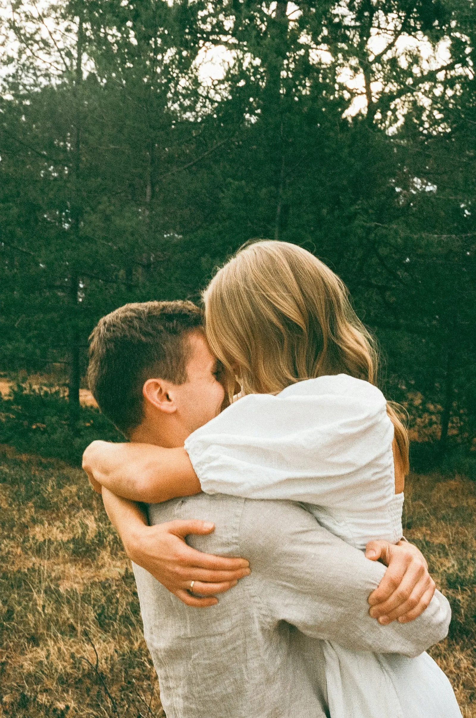 A couple in linen embrace in a grassy clearing with pine trees behind them.