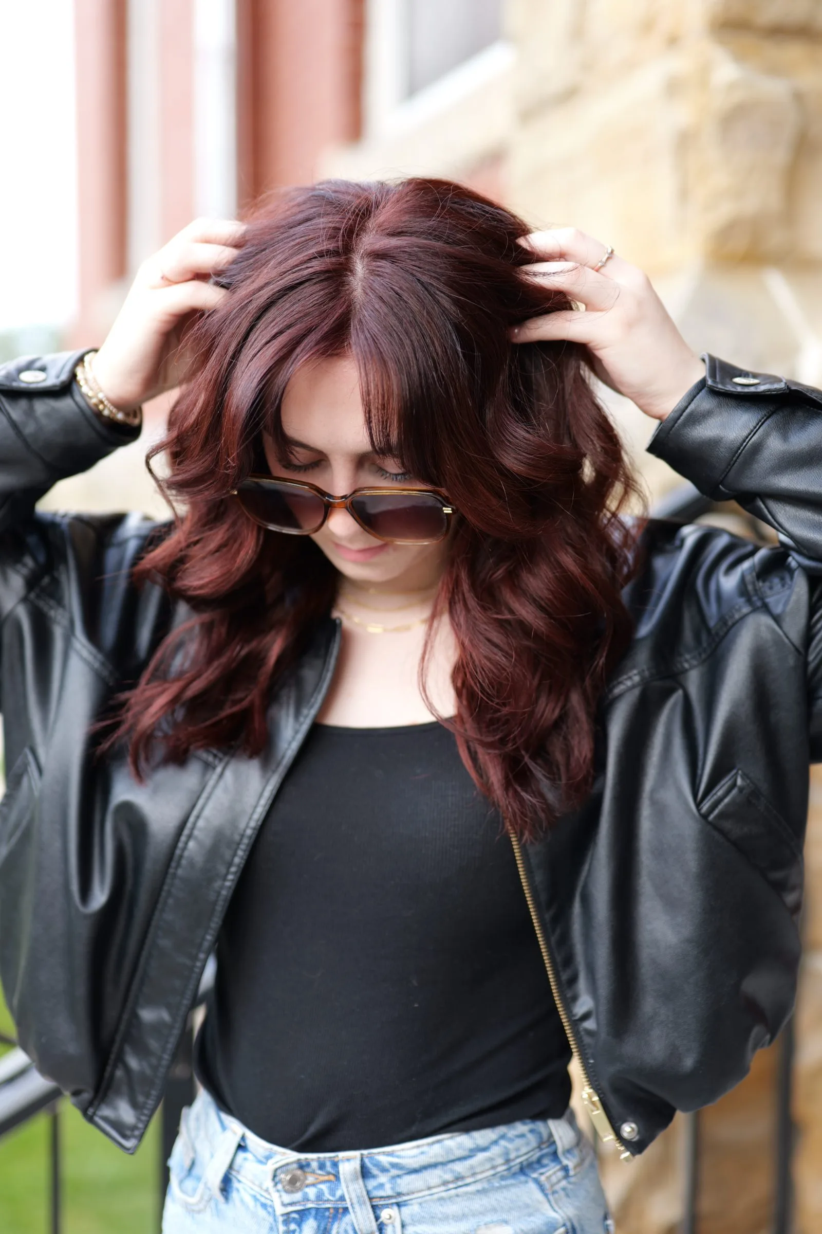 A woman with auburn hair in a leather jacket, hands in her hair, looking down.